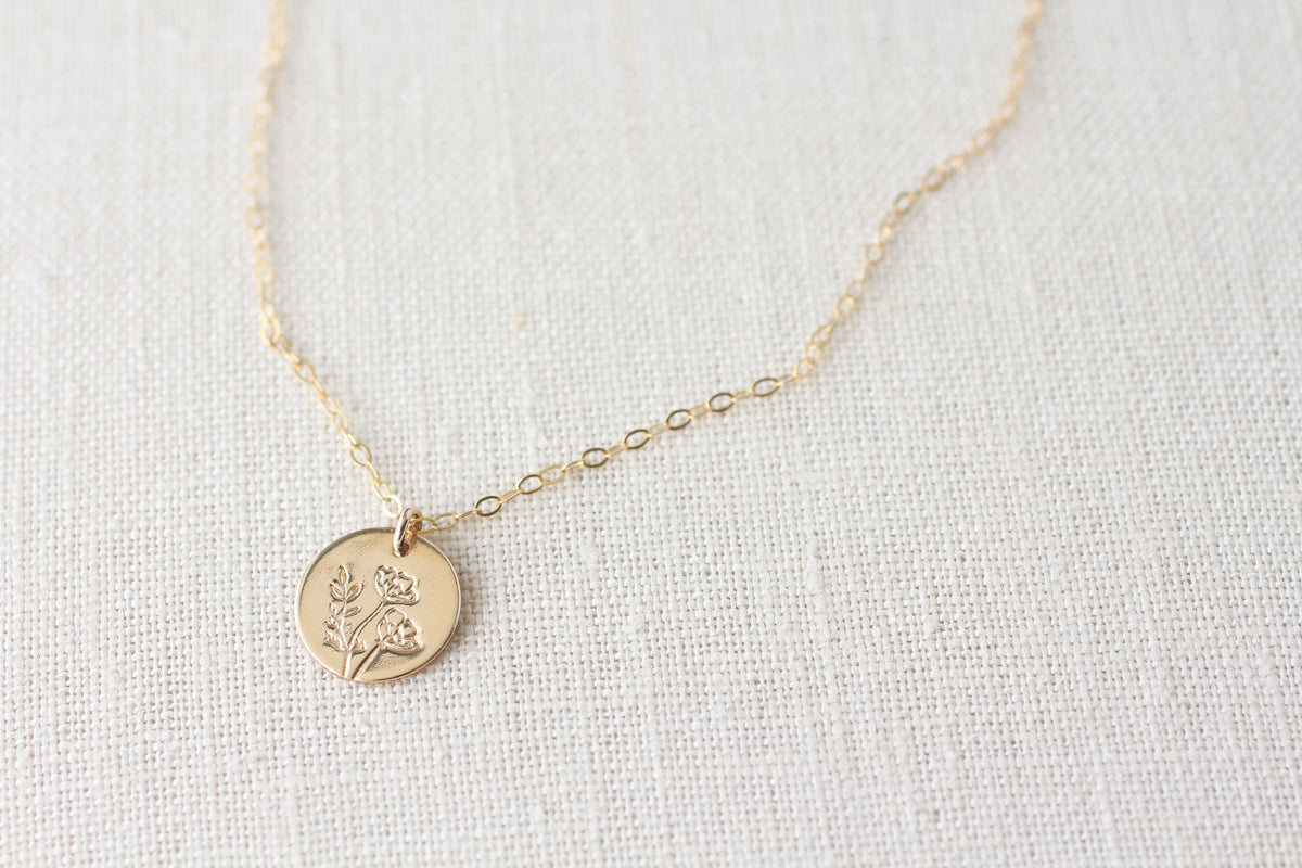 A delicate gold chain necklace with a small round gold coin pendant engraved with a floral design, displayed on a textured beige linen fabric background.