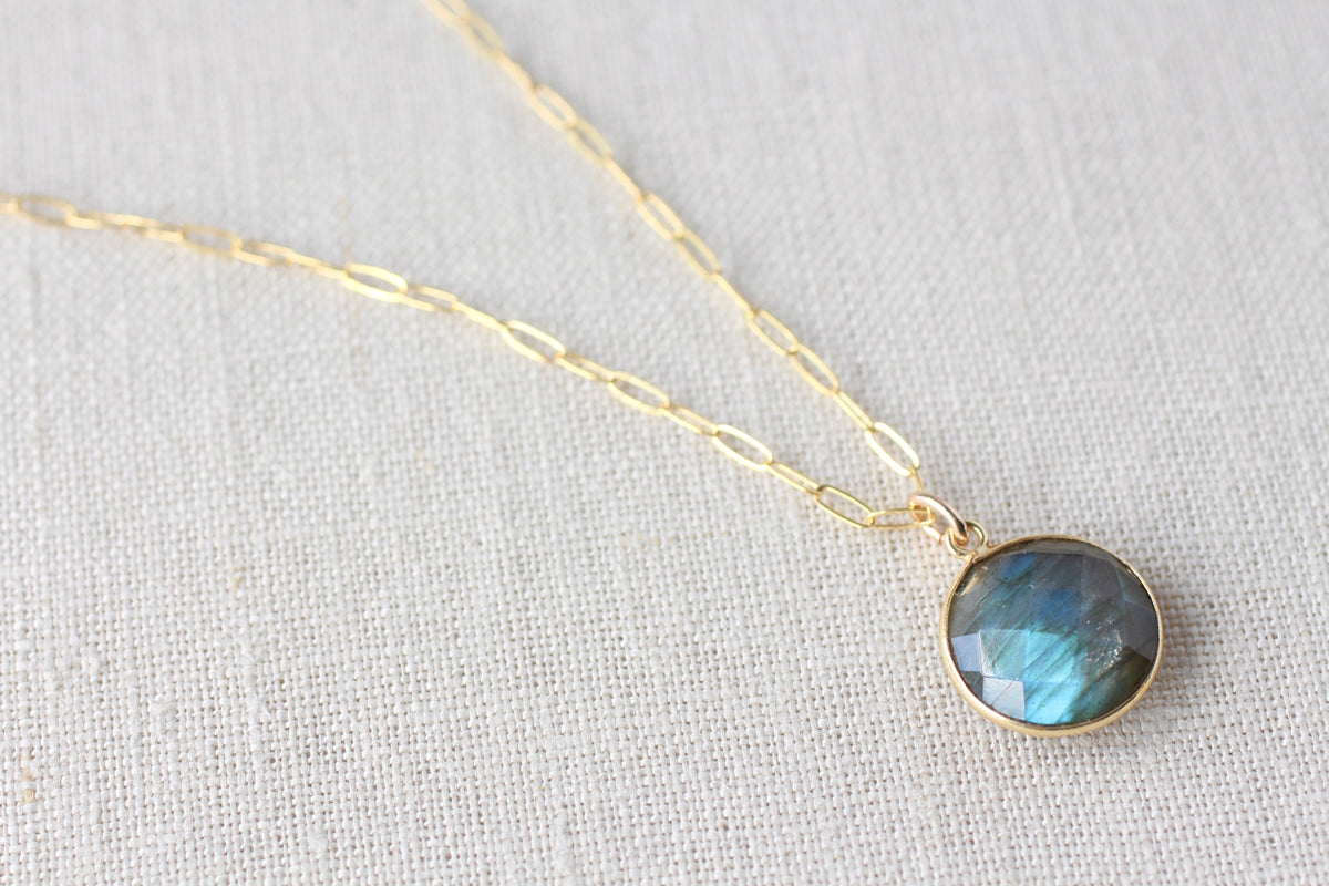 A gold paperclip chain necklace with a round bezel-set blue-green labradorite gemstone pendant, displayed on a textured off-white linen background.