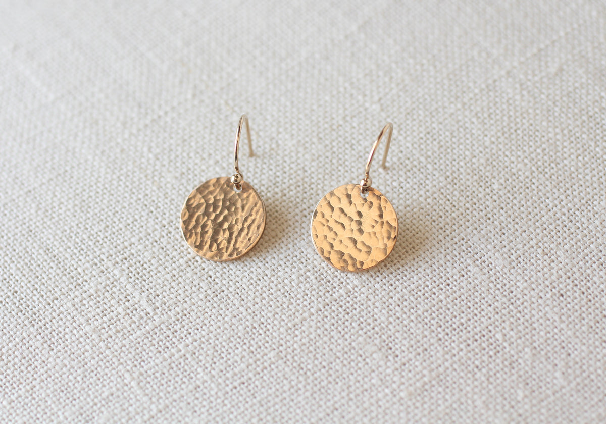 A pair of hammered gold round drop earrings with fishhook wires, displayed on a textured off-white linen background.