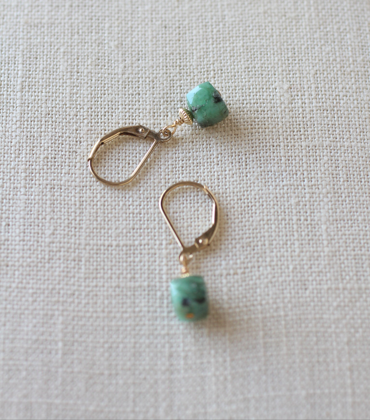 A pair of gold-toned leverback earrings with small cube green and black stone drops, displayed on a textured beige linen fabric background.