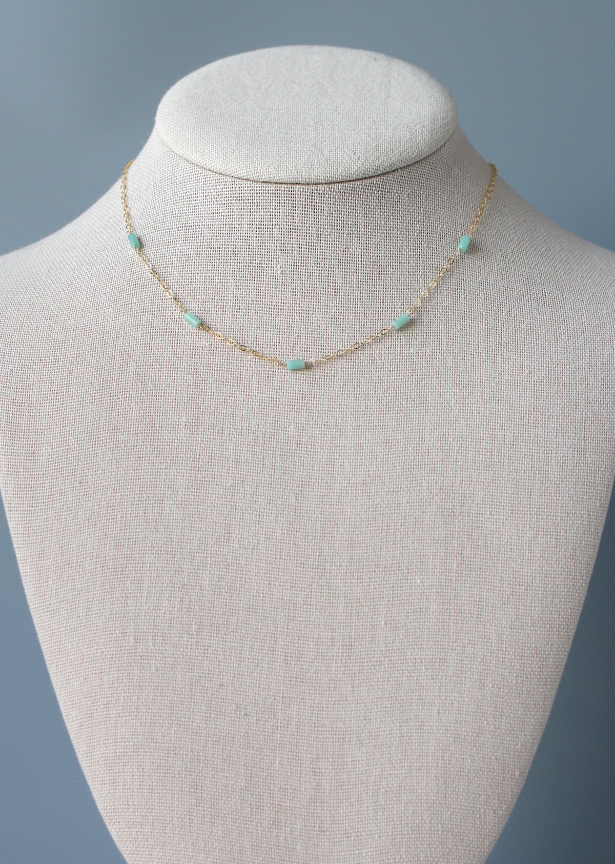 A delicate gold chain necklace with small mint green beads, displayed on a textured beige fabric jewelry bust against a soft blue-gray background.