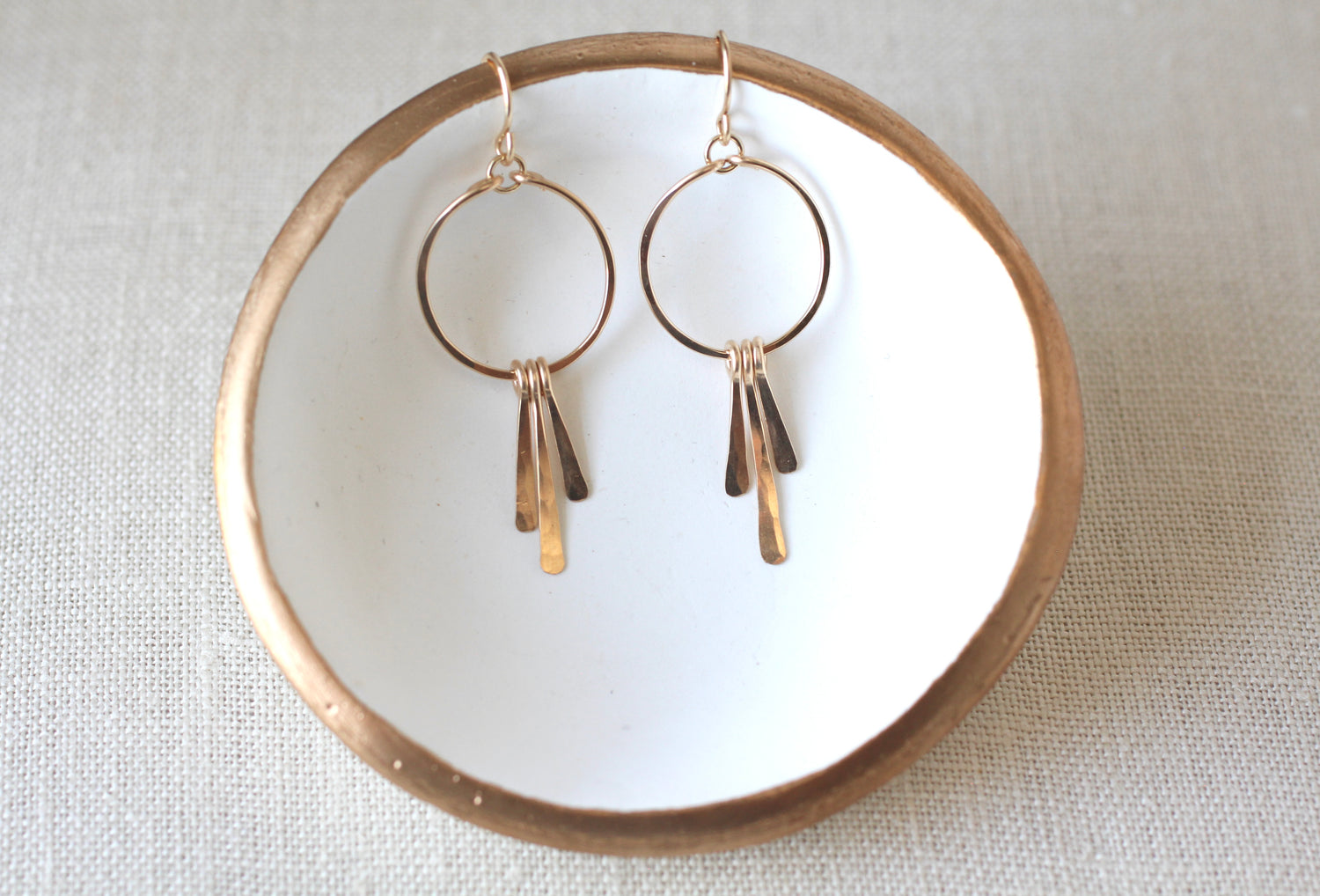 A pair of gold-toned dangle earrings with circular hoops and three dangling bar charms, displayed on a white ceramic dish with a gold rim over a light beige fabric background.
