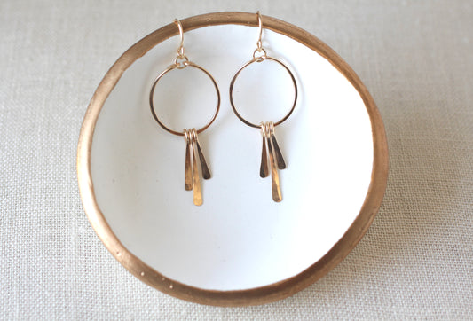 A pair of gold-toned dangle earrings with circular hoops and three dangling bar charms, displayed on a white ceramic dish with a gold rim over a light beige fabric background.