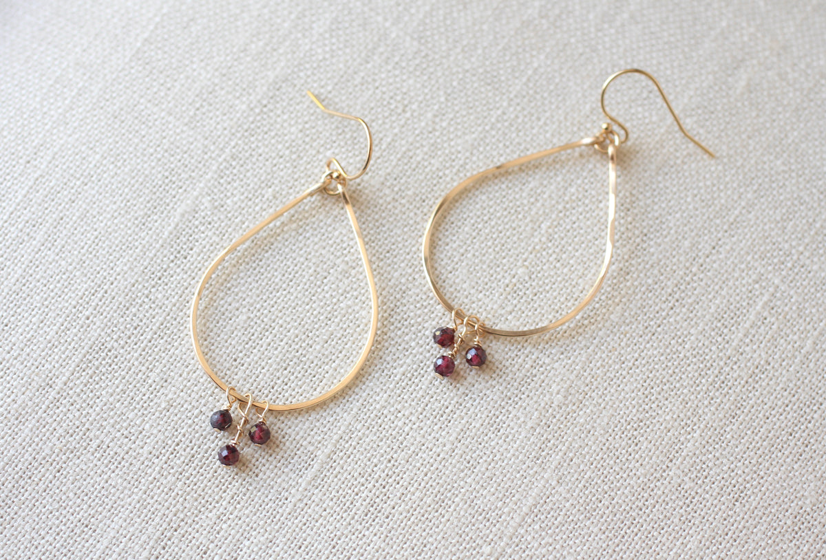 A pair of gold teardrop hoop earrings with small dangling burgundy gemstone clusters, displayed on a textured off-white linen background.