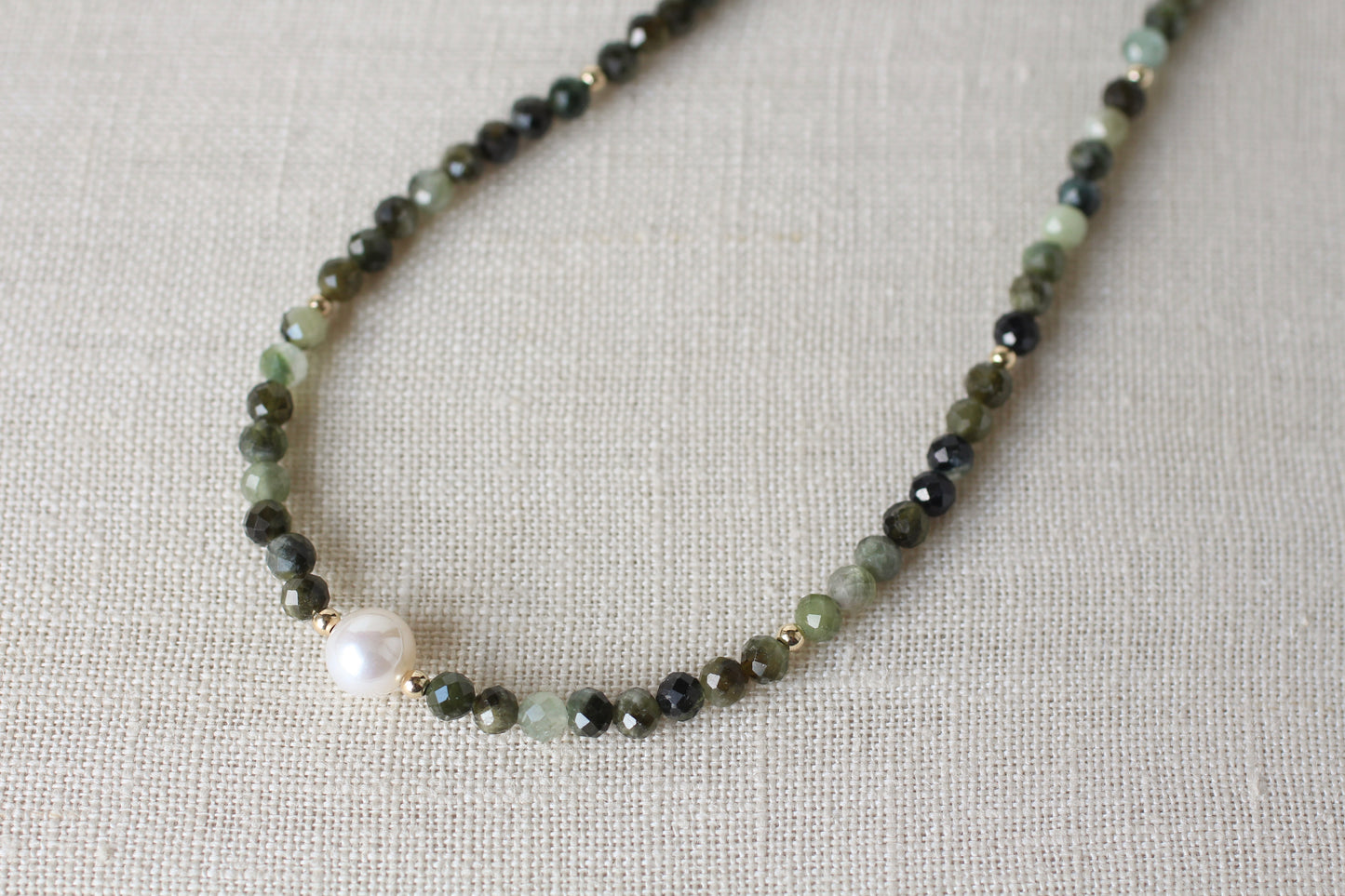 A beaded necklace featuring faceted dark green and light green beads, small gold spacer rings, and a single prominent white pearl bead, displayed on a textured beige linen background.