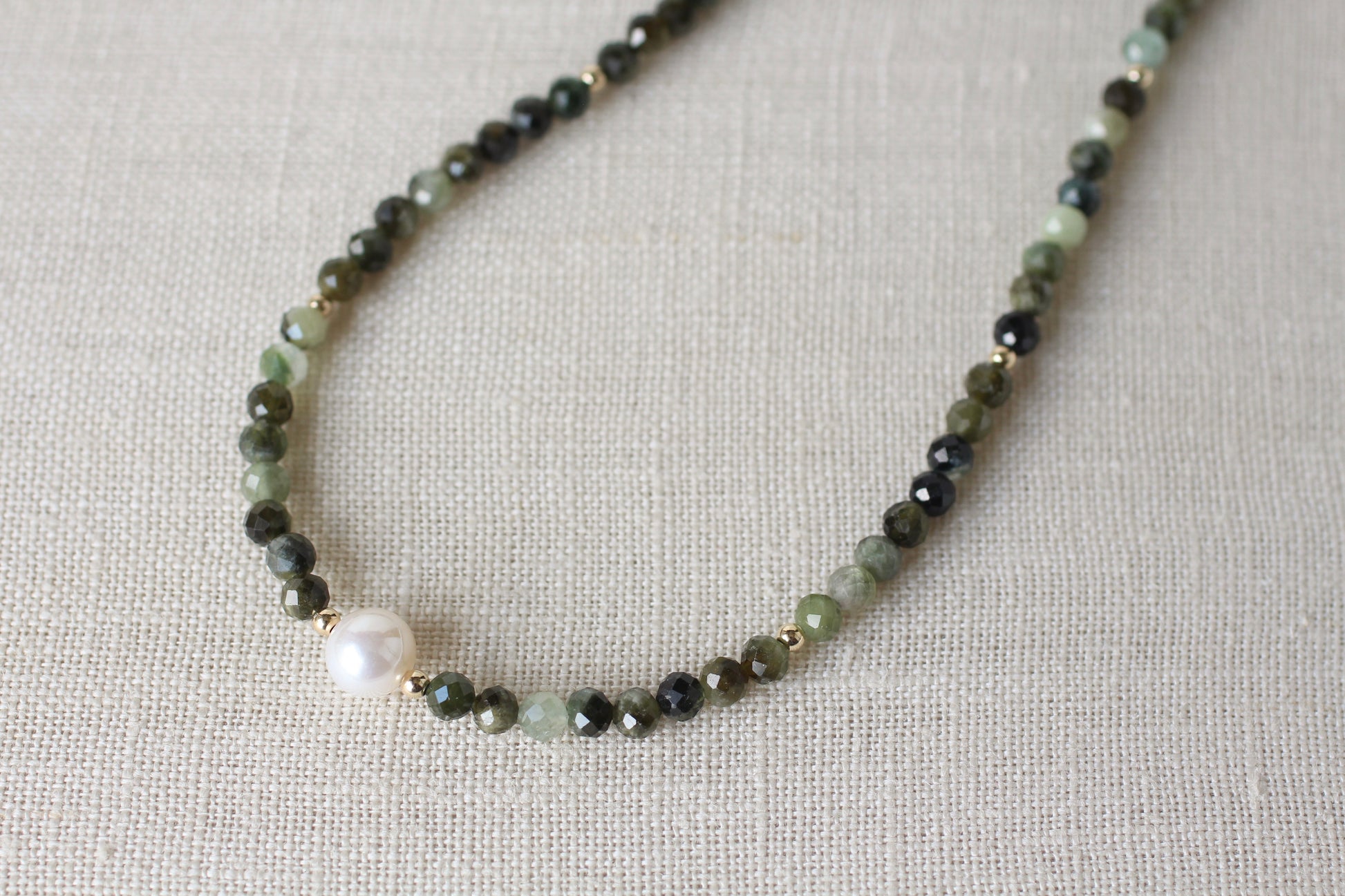 A beaded necklace featuring faceted dark green and light green beads, small gold spacer rings, and a single prominent white pearl bead, displayed on a textured beige linen background.