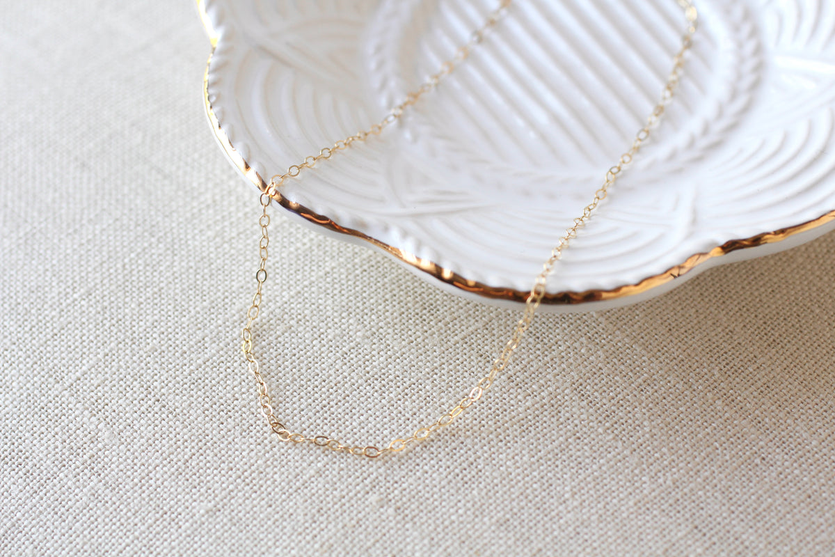 A delicate gold chain necklace draped over a white textured ceramic plate with a gold-trimmed edge, set on a beige linen fabric background.