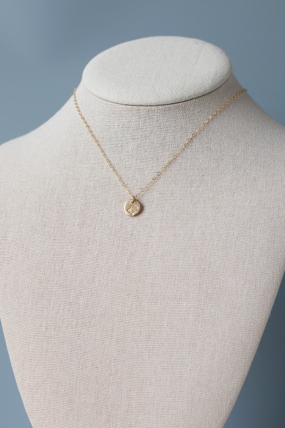 A delicate gold chain necklace with a small, textured circular pendant displayed on a cream-colored fabric jewelry bust against a soft blue-gray background.