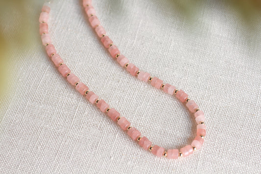 A delicate pink beaded necklace with small gold spacer beads, arranged in a loop on a textured off-white linen fabric background.