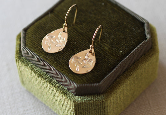 A pair of gold teardrop dangle earrings with engraved leaf patterns, displayed on a green velvet jewelry box.