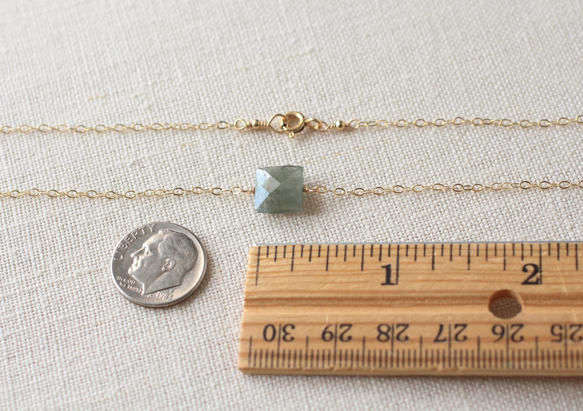 A delicate gold chain necklace with a small square-cut green gemstone pendant, displayed on a textured beige fabric alongside a US dime and a wooden ruler for scale.