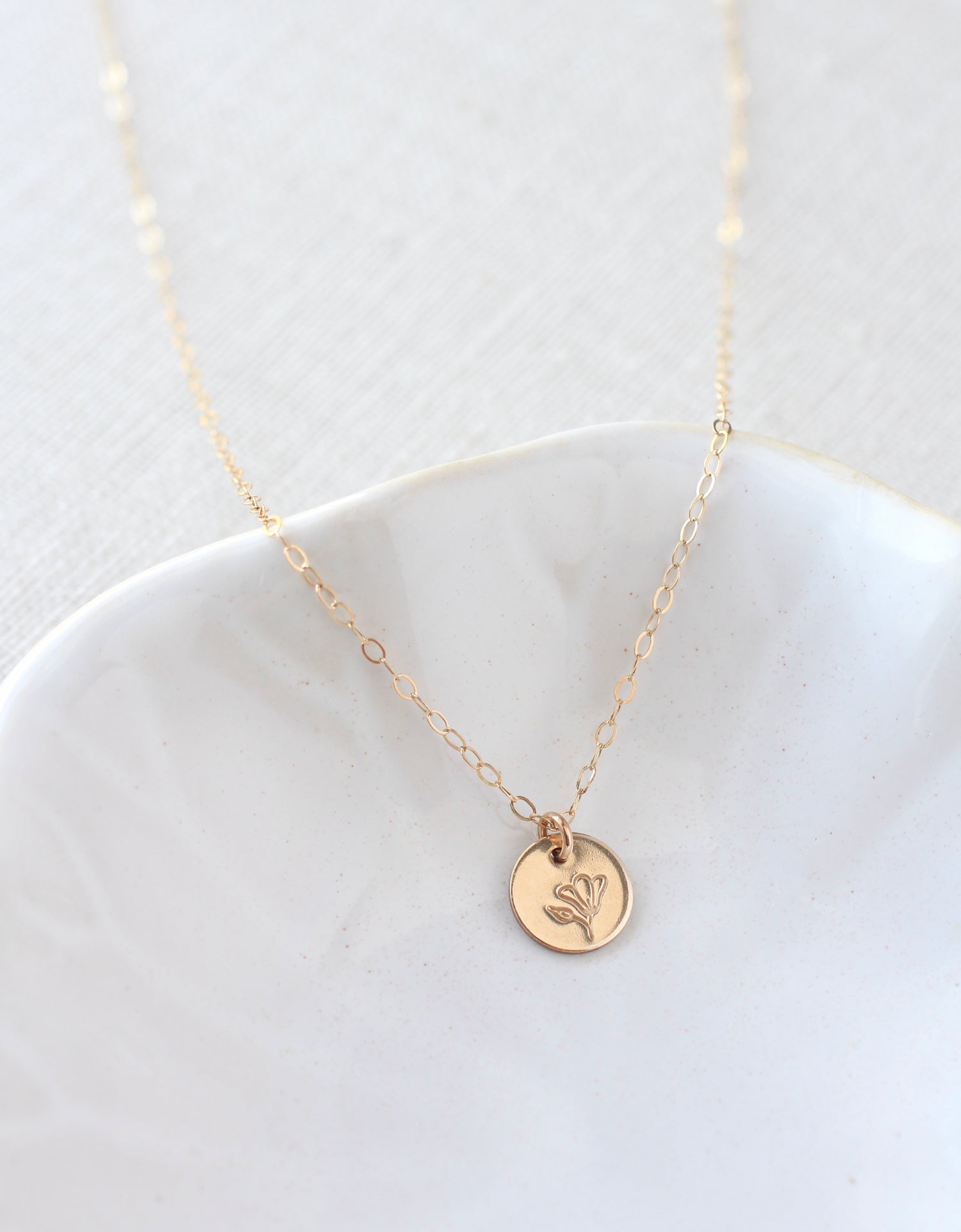 A delicate gold chain necklace with a small round gold disc pendant featuring an engraved floral design, displayed on a white ceramic dish against a light textured background.