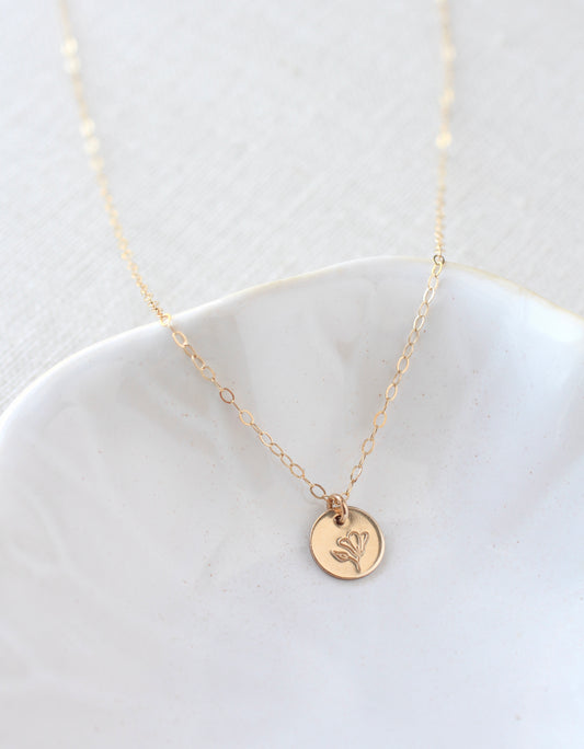 A delicate gold chain necklace with a small round gold disc pendant featuring an engraved floral design, displayed on a white ceramic dish against a light textured background.