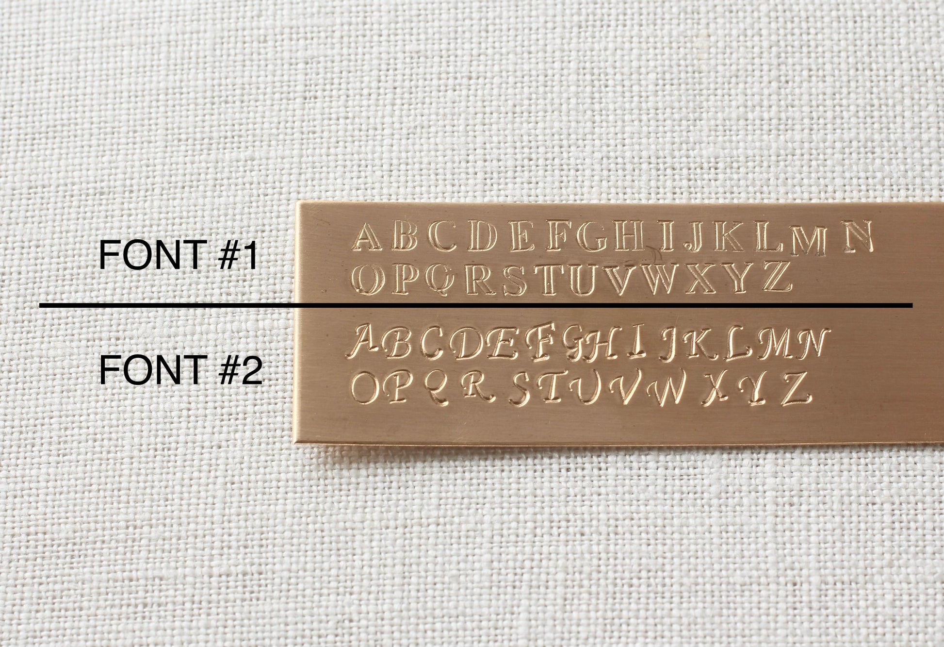 A rectangular brass-colored metal sample plate on a textured off-white fabric background, showing two font styles labeled FONT #1 and FONT #2. The top row of letters in FONT #1 is cut off, while FONT #2 shows the full alphabet from A to Z in raised, polished lettering.