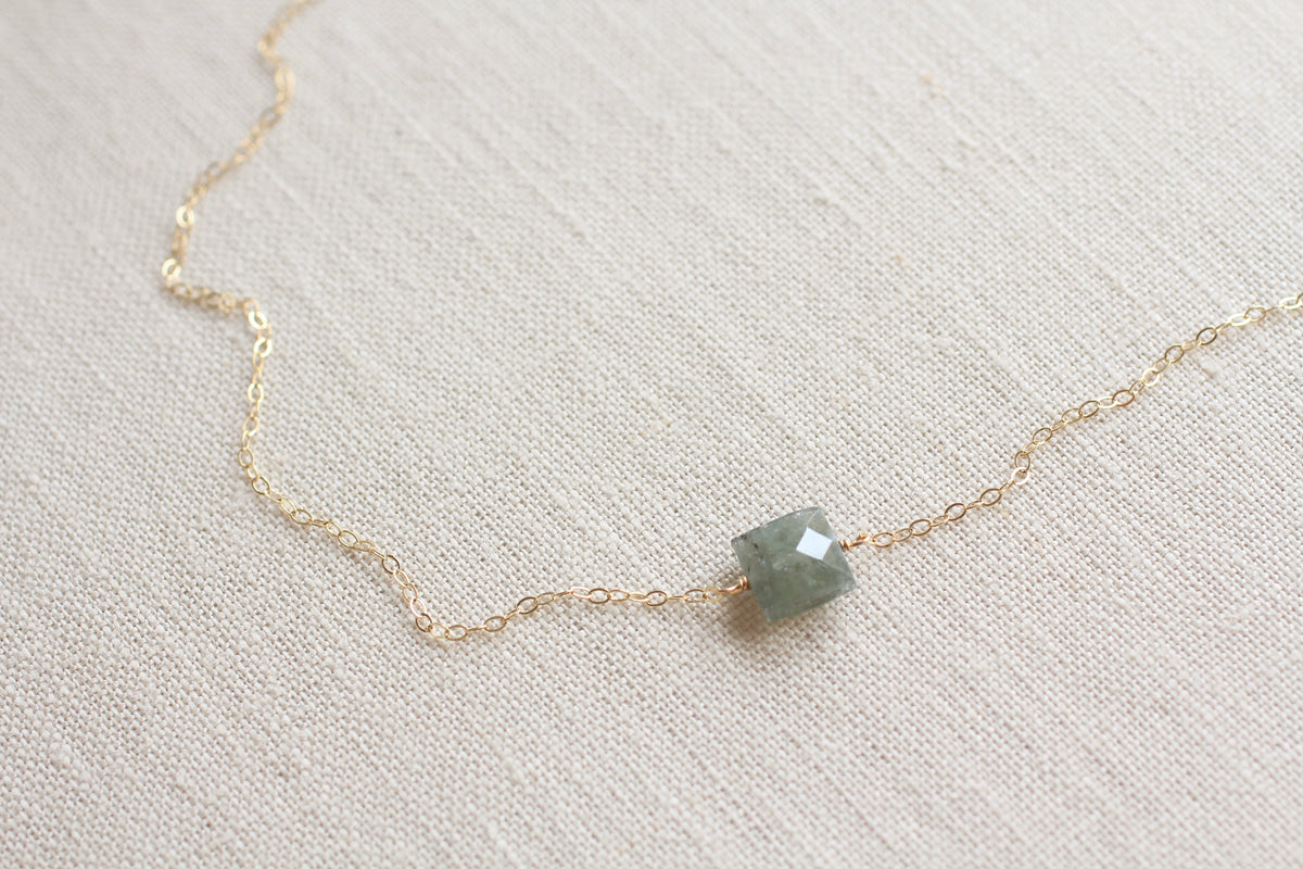 A delicate gold chain necklace featuring a small, faceted green gemstone pendant, displayed on a textured beige linen fabric background.