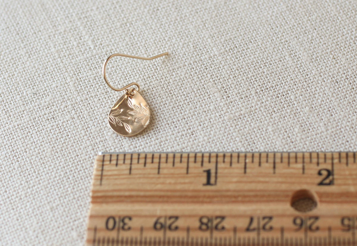 A small gold teardrop-shaped dangle earring with a leaf motif, shown next to a wooden ruler on a textured beige fabric background.