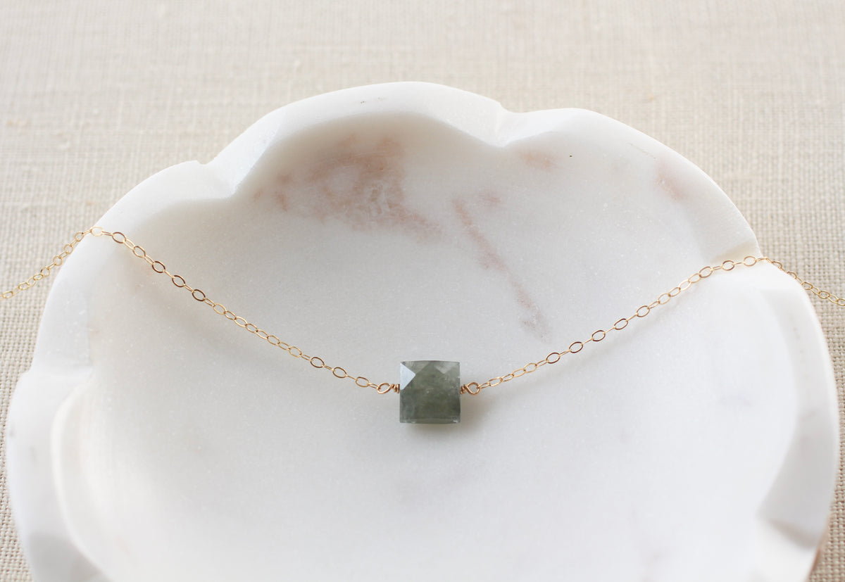 A delicate gold chain necklace with a small, square, faceted gray-green stone pendant, displayed on a white scalloped marble dish over a beige linen background.