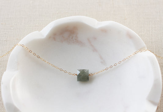 A delicate gold chain necklace with a small, square, faceted gray-green stone pendant, displayed on a white scalloped marble dish over a beige linen background.