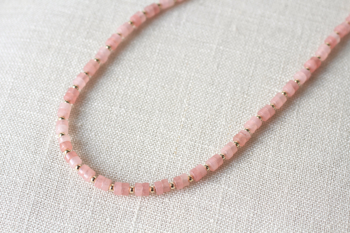 A close-up of a pink faceted bead necklace with small gold spacer beads, laid on a textured off-white linen background.