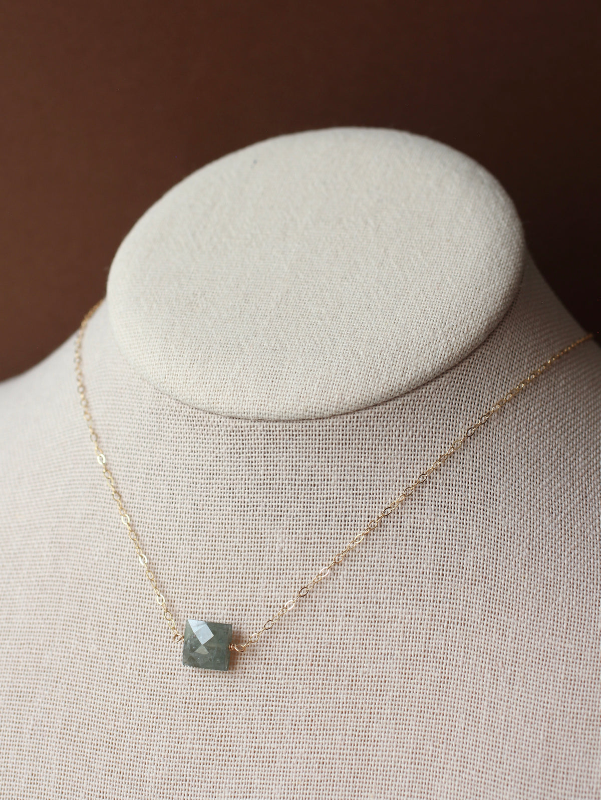 A delicate gold chain necklace with a small square faceted gray-green gemstone pendant, displayed on a cream-colored fabric jewelry bust against a dark brown background.