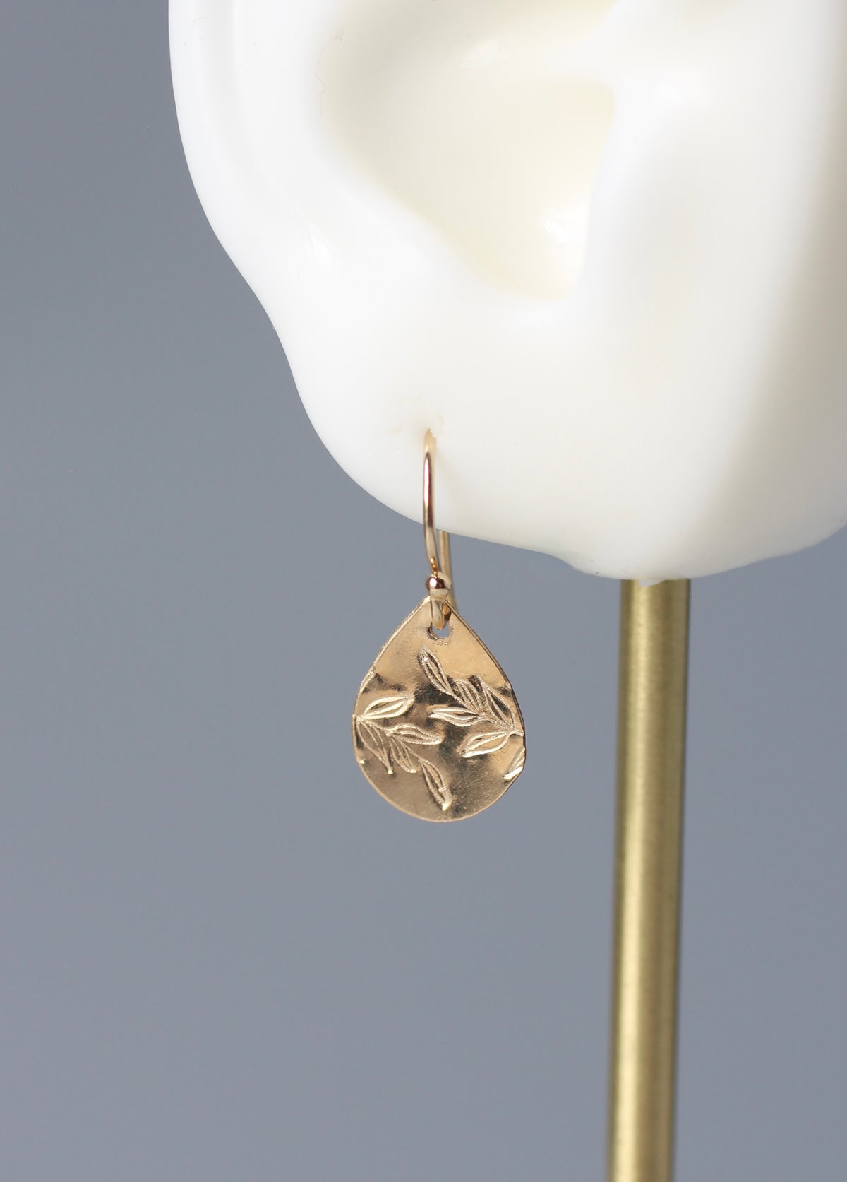 A gold teardrop-shaped drop earring with a stamped leaf pattern, displayed on a white ear-shaped jewelry stand against a gray background.