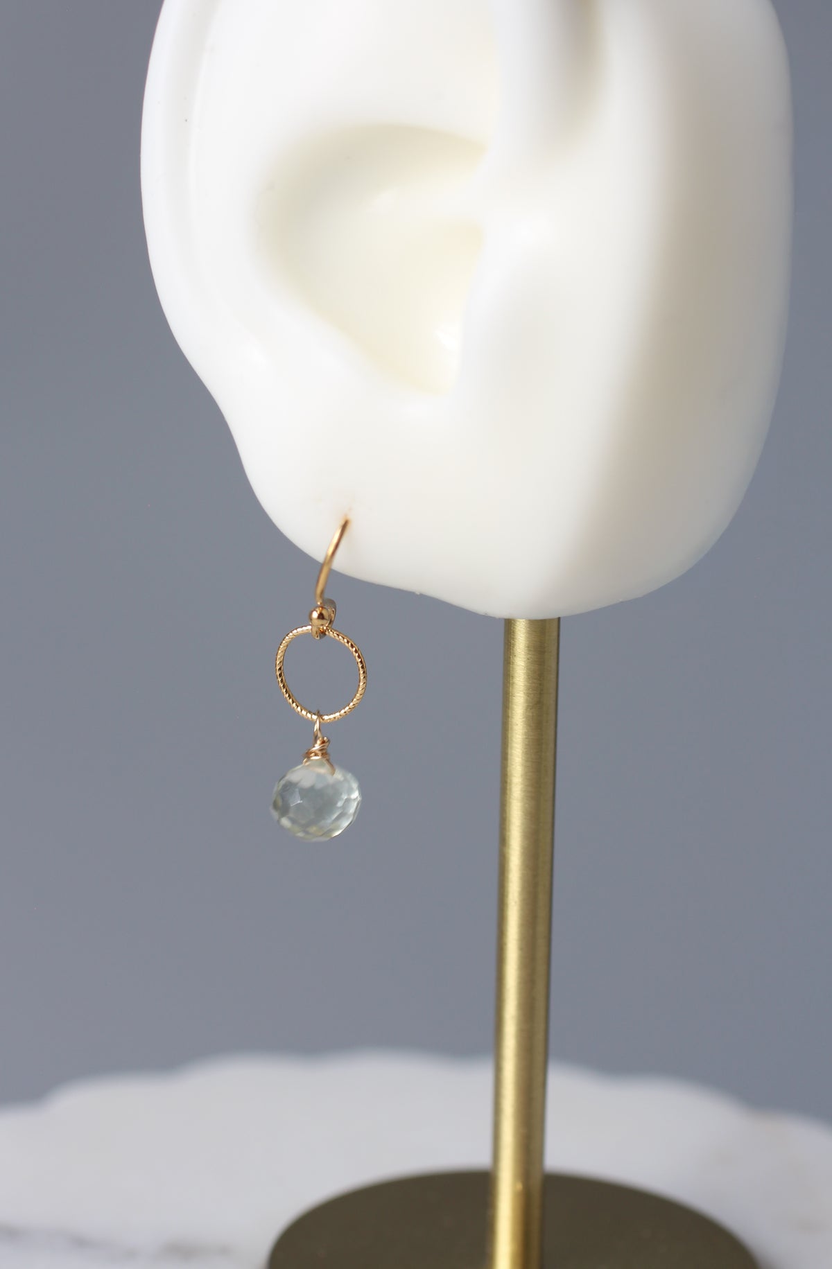 A delicate gold hoop earring with a small faceted clear gemstone dangle, displayed on a white ear mannequin head on a gold stand against a soft gray background.